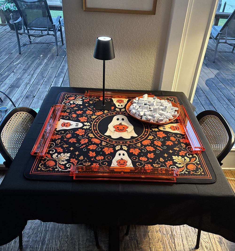 A square mahjong table set for Halloween: a black cloth, a ghost and pumpkin mat, four orange racks, a small black lamp and a bowl of tiles