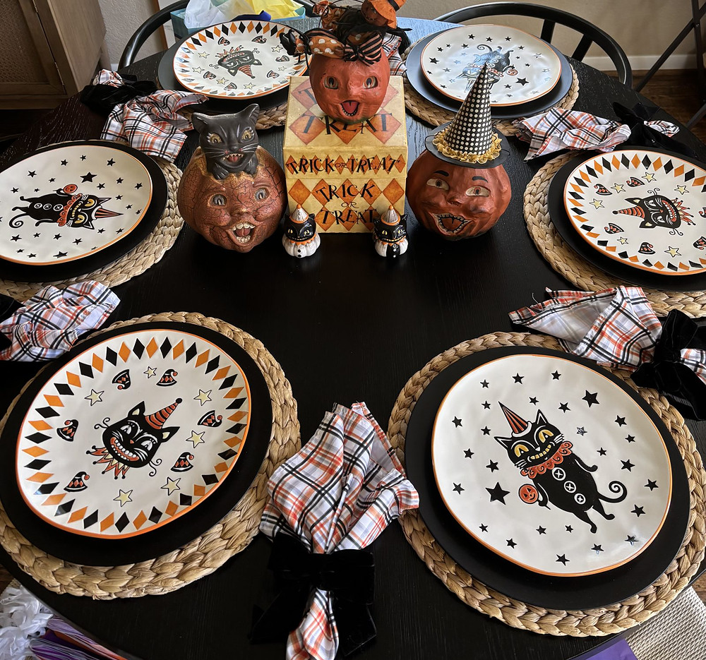 A round dinner table set for Halloween with black cat plates on woven chargers, plaid napkins tied in black, and pumpkin head figures around a Trick or Treat box